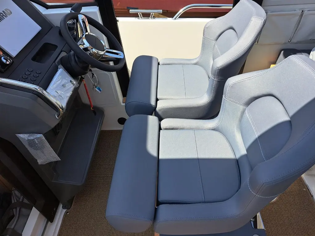 Boat cockpit featuring a grey two-person seat and a steering wheel, with controls and a display panel visible.